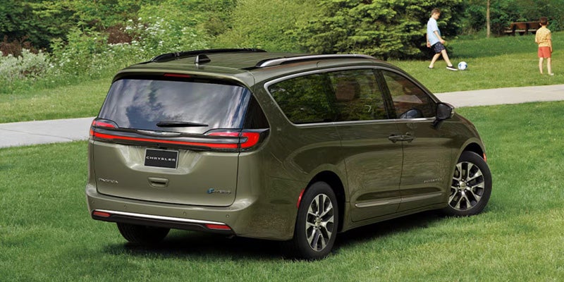 Green minivan parked on grass near a park with trees and people in the background.