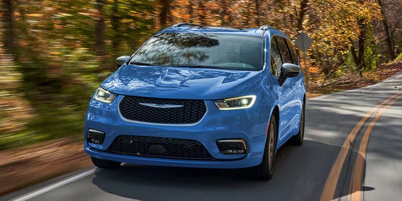 Blue minivan driving on a winding road surrounded by trees, shown in front view