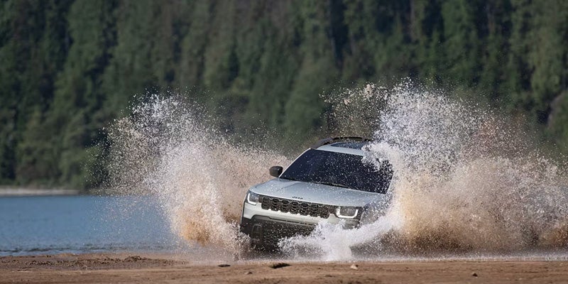 SUV driving through shallow water, creating large splashes with a forested background behind it