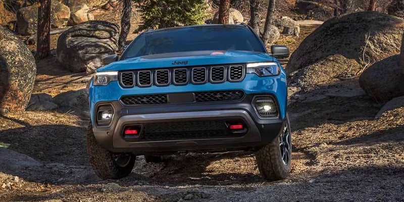 Blue SUV driving over rocky terrain in a forested area, shown in front view