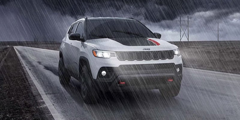 White Jeep SUV driving on a wet road in heavy rain, with headlights on and water spray visible