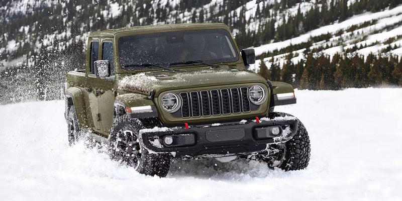 Green Jeep pickup driving through snow-covered terrain, creating snow spray around the vehicle