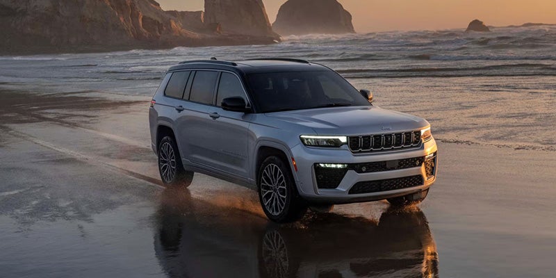 White SUV driving along a sandy beach at sunset with ocean waves, rocky cliffs