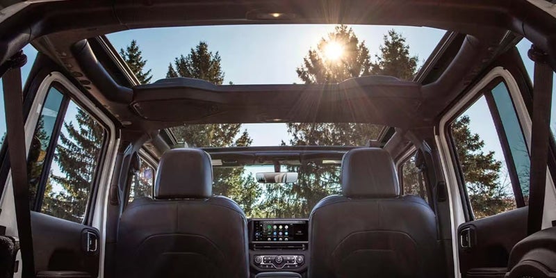 Interior view of a Jeep Wrangler with an open panoramic roof and sunlight shining through trees