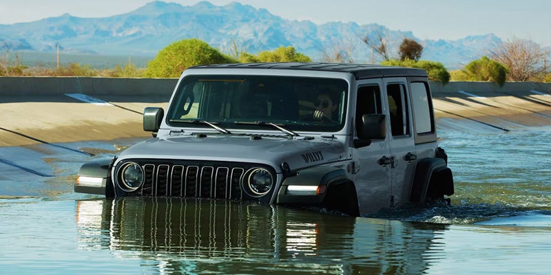 Gray Jeep Wrangler partially submerged while driving through shallow water near mountains