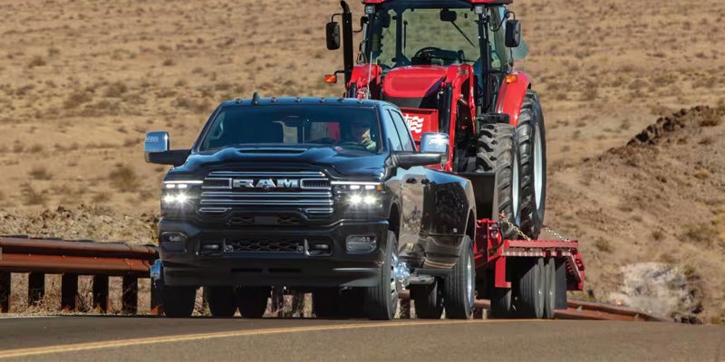 Black RAM pickup truck towing a tractor on a trailer along a desert road