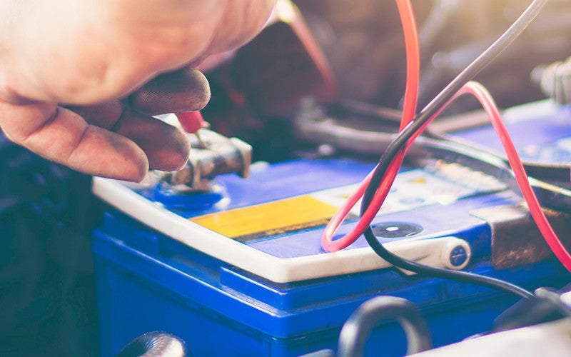 A person checking a blue car battery with red and black wires, their fingers appearing from the top left.